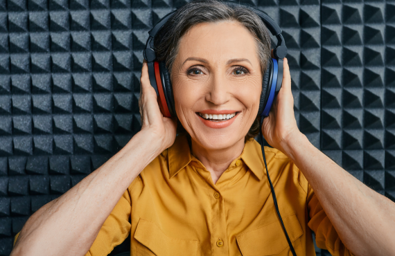 A smiling older woman in a mustard yellow shirt wearing large over-ear headphones, holding them with both hands, in a soundproof studio with acoustic foam panels in the background.